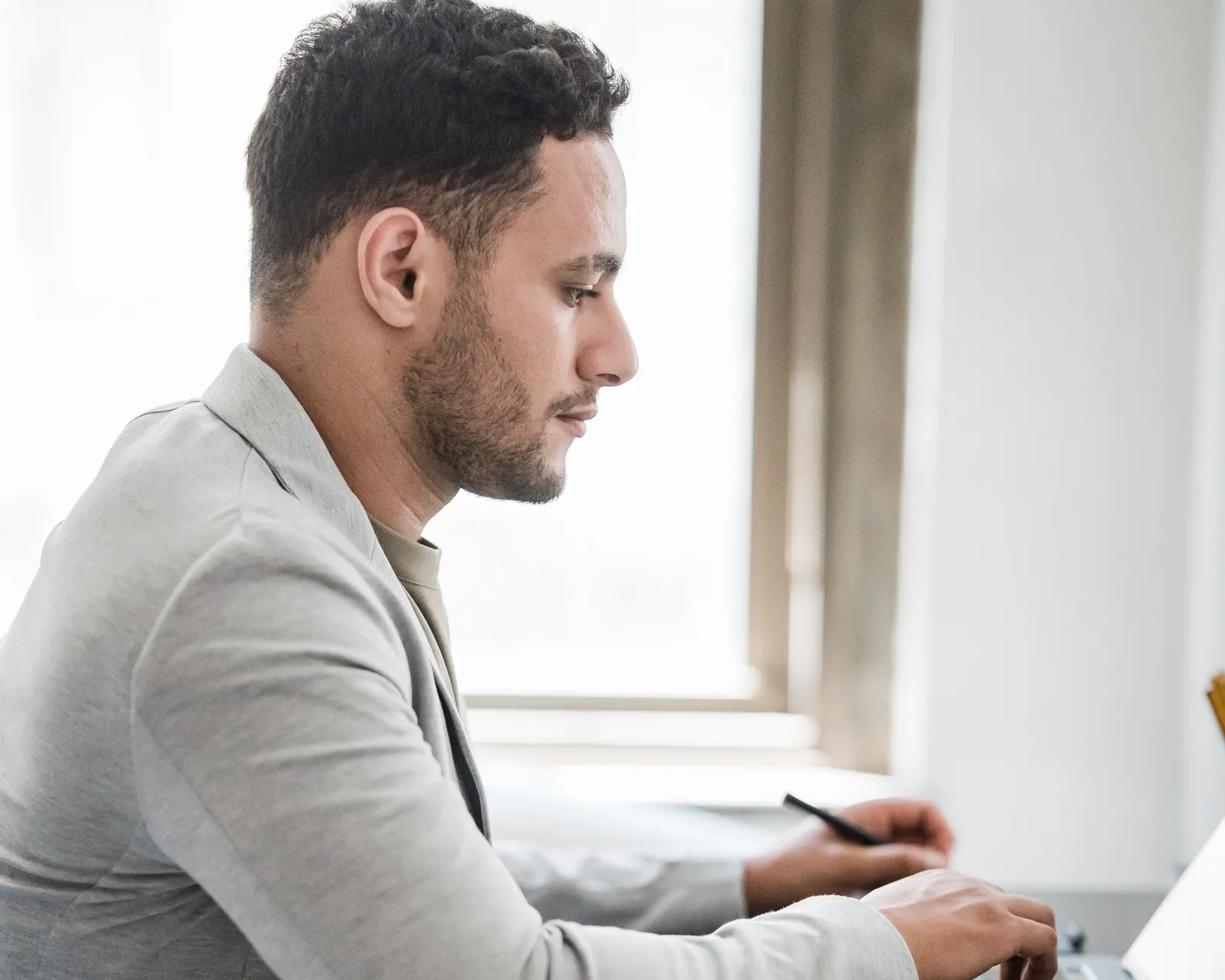 A man at a desk by a window, pen in hand, working at a laptop.