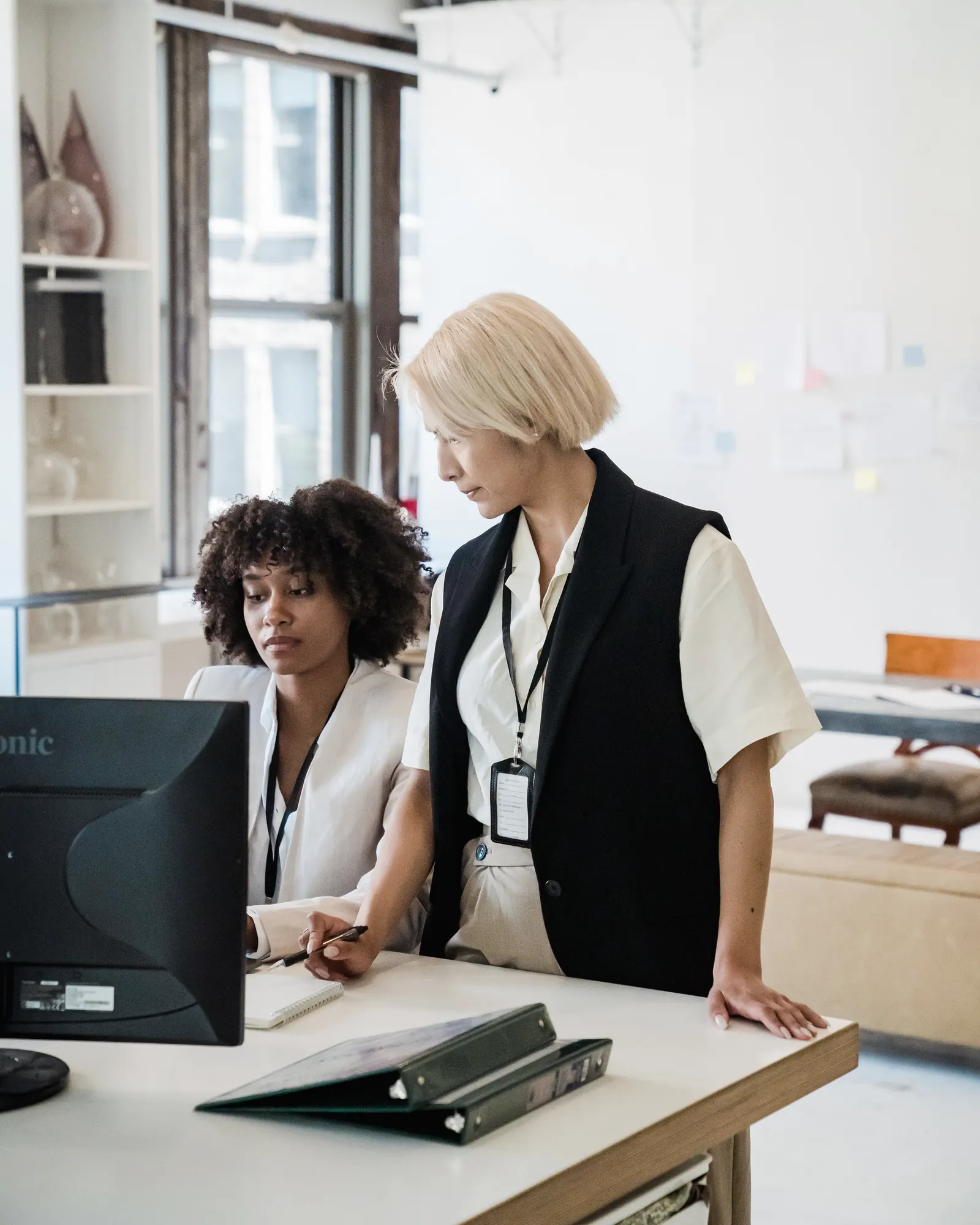 Two colleagues at a desk, one at a monitor, the other leaning on the desk beside her.
