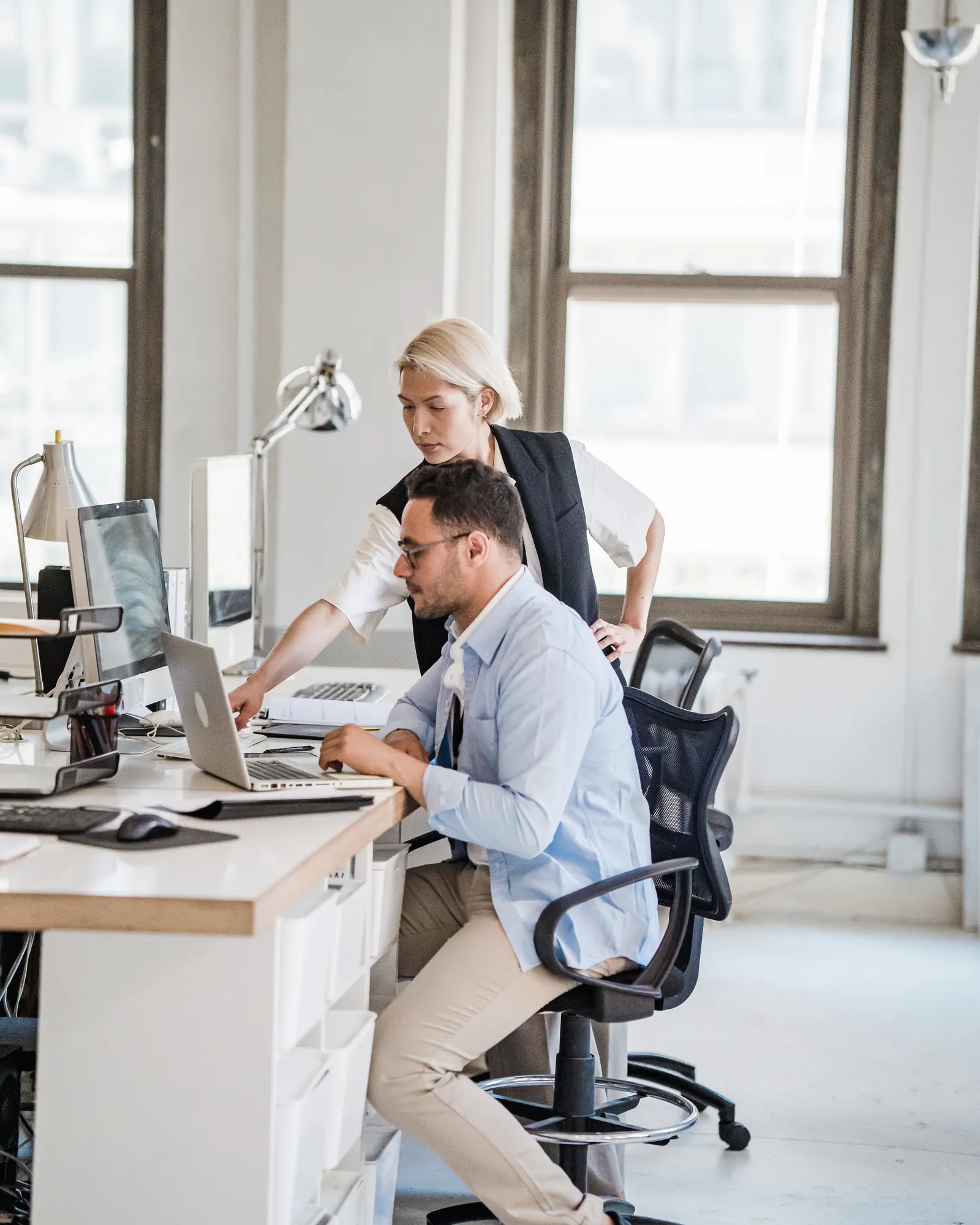 A bright open office: one colleague at his desk, another leaning in to look at his screen.