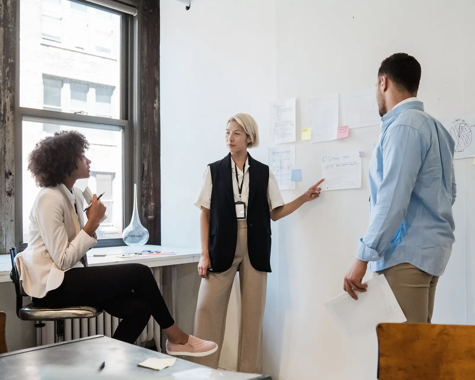 Three colleagues at a wall of pinned notes by a tall window; one points to a sheet as she talks it through.