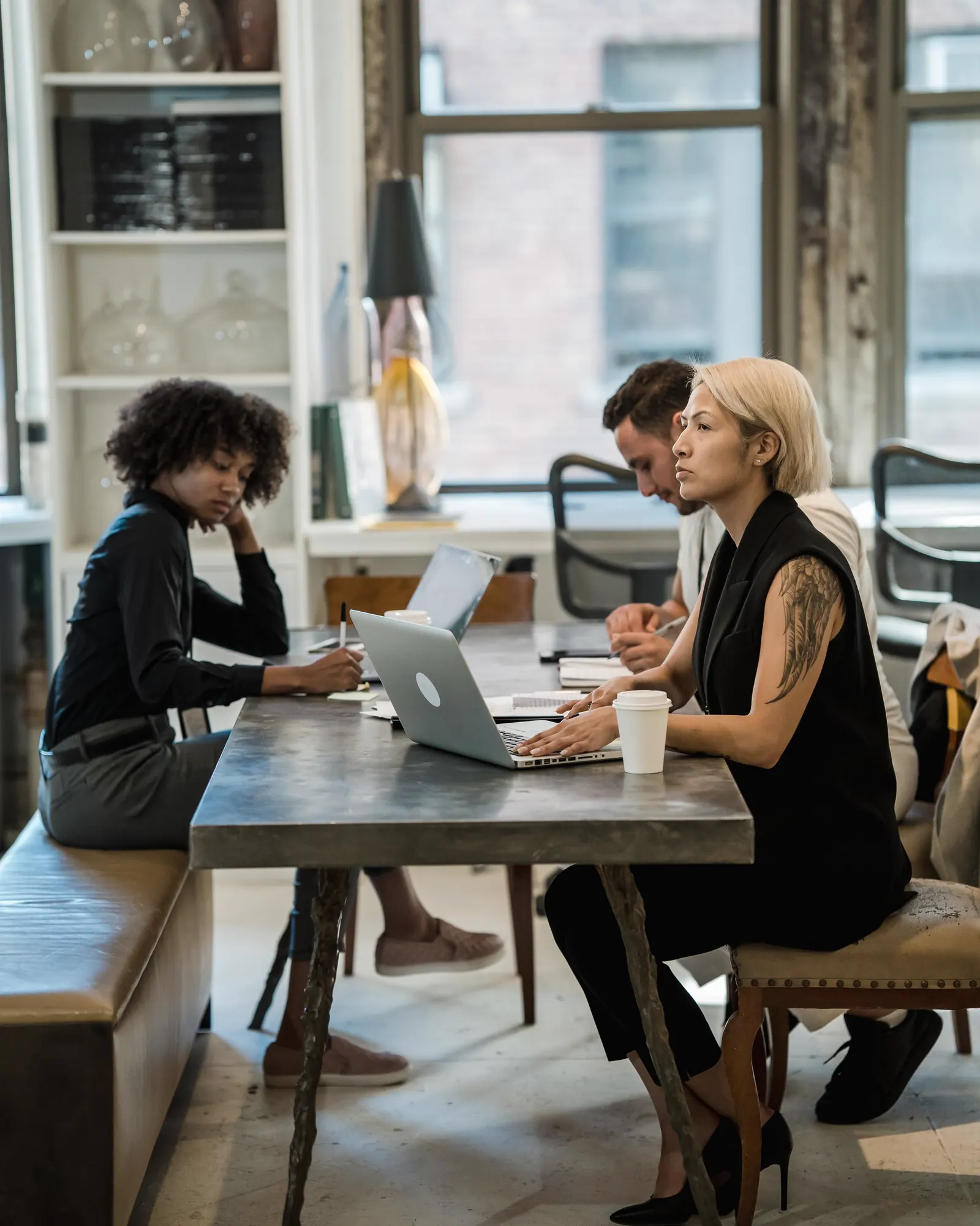 A team at a long table by tall windows, laptops open, coffee between them.