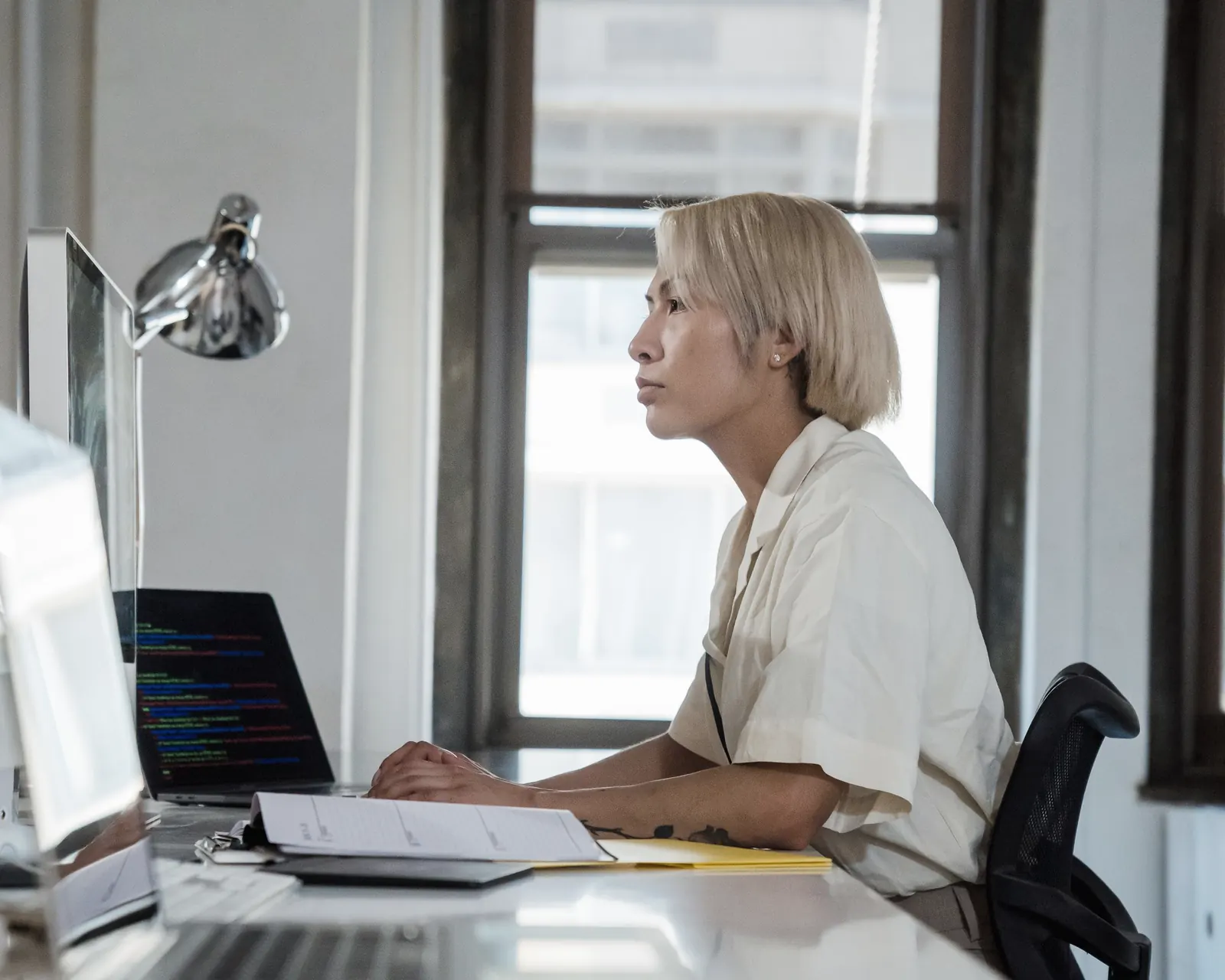 A woman at a long desk by the window, working between a laptop and a monitor.