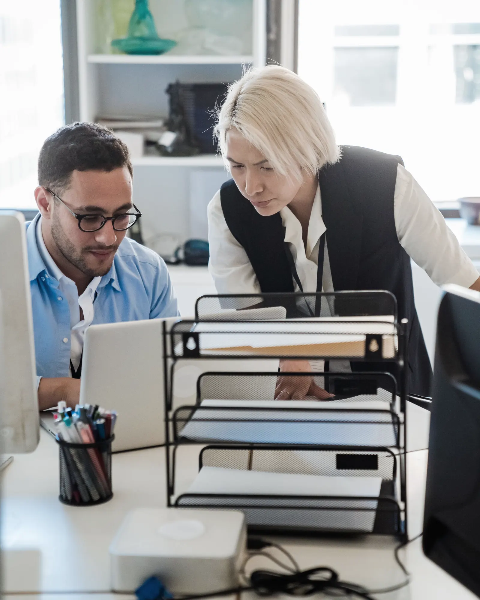 A colleague leans over a desk to look at a laptop with the man working at it.